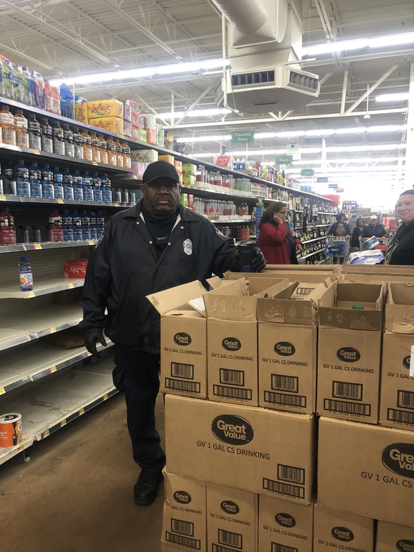 A police officer that is standing in an isle in a supermarket. Next to him are more than a dozen of cardboard boxes that contain Great Value water. Behind the man in the isle are shoppers browzing the isle.