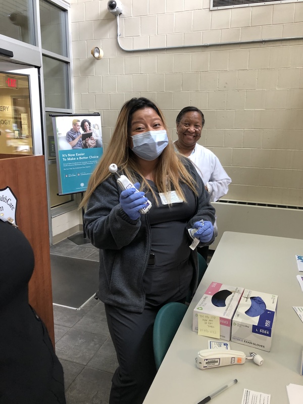 A healthcare worker wearing sterile gloves and a facemask holding a thermometer. Another person standing behind them without a facemask smiling. Both standing in front of a table with sterile gloves and another thermometer on top of it.