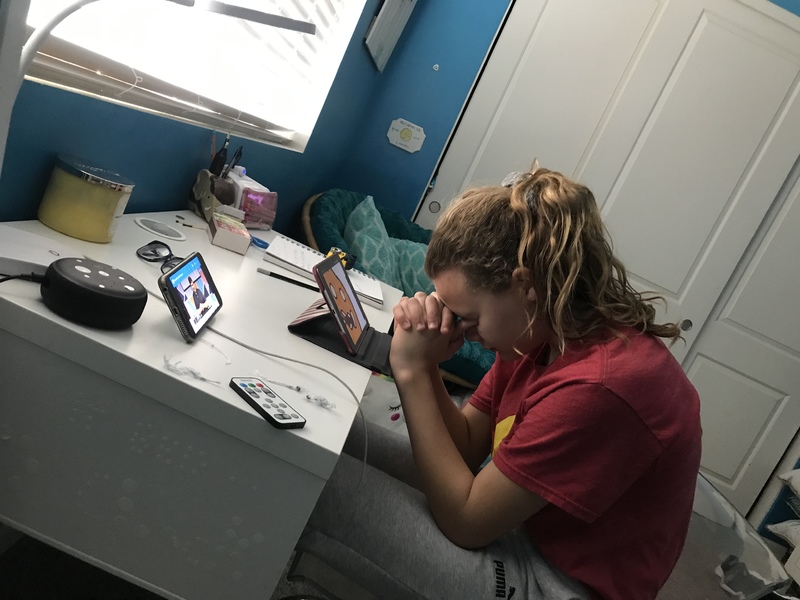 A person seating at a white desk wearing sweatpants and a red shirt. The person has their hands folded and placed their head against their hands. They are praying. A church service is being played on a propped up iPhone on the desk in front of them. 