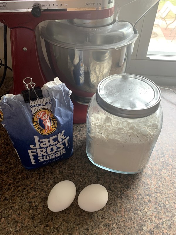 A red Artisan Kitchen Aid, a mason jar with a silver lid filled with white flour, two white eggs, and a blue Jack Frost granulated pure cane sugar bag on a speckled countertop surface.
