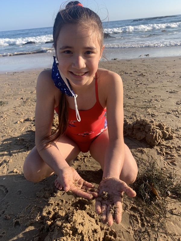 This is a picture of a young girl in a red swimsuit playing in the sand at the beach, and smiling into the camera. A face mask hangs off of one of her ears.