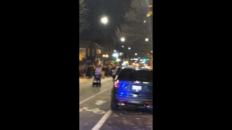 Blurry image of a street at night.  There is a small, blue car in the foreground and pedestrians walking on the street.