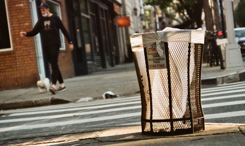 A photo of a trash can with a man in a lowered mask in the background.