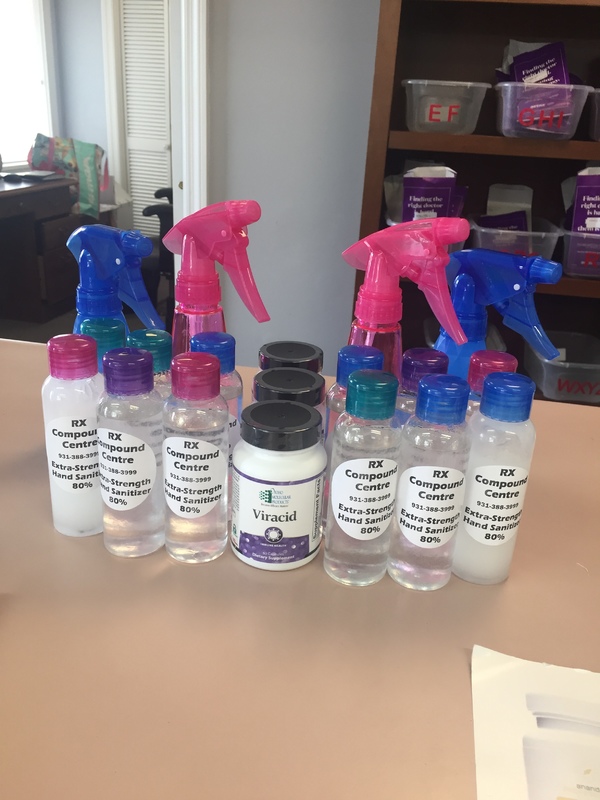 A counter has various bottles of hand sanitizer with labels on them that say: RX Compound Centre. Near the hand sanitizer bottles are also bottles of Viracid.