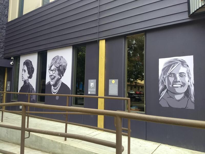 This is a picture taken of a series of murals that are painted on the side of a building. Various women in different periods of clothing are depicted in black and white.