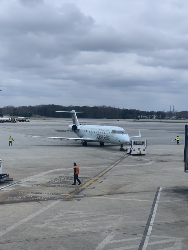 A white plane is parked outside. There are various people wearing orange and yellow safety vests walking around the plane.