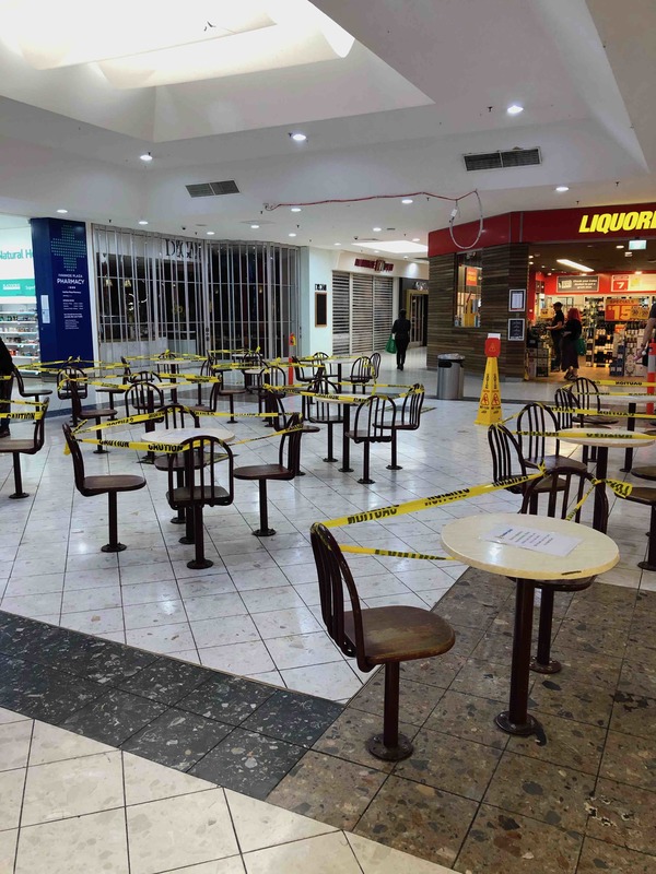 Inside of a shopping mall has caution yellow tape wrapped around seats and tables to block them off from people being able to use them.