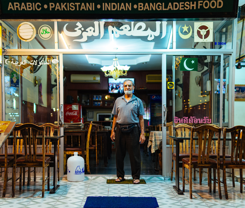 A shop owner stands outside his restaurant. The shop owner is wearing a grey polo with black pants, around his waist is a black fanny pack. He is wearing brown sandals. Above him is the sign to his restaurant that says: ARABIC PAKISTANI INDIAN BANGLADESH FOOD. To the right and left of him are wooden table and chairs for outdoor dining.