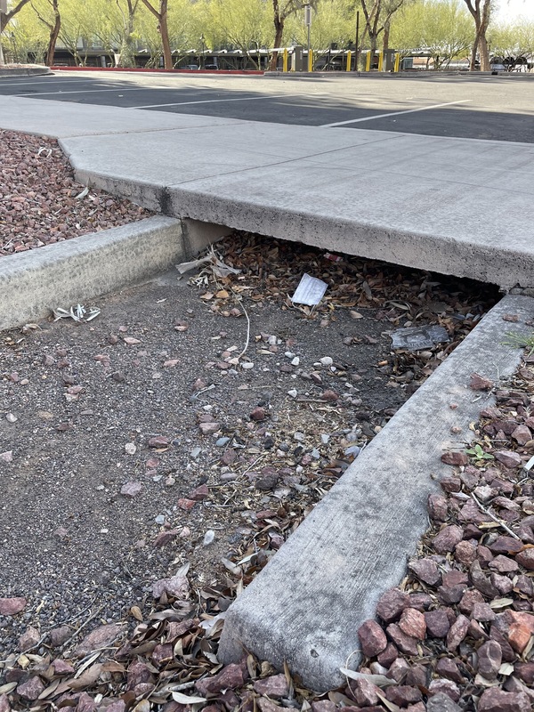 This is a picture of a face mask that has been discarded in the dirt below a drainage area. A concrete walkway extends above it.