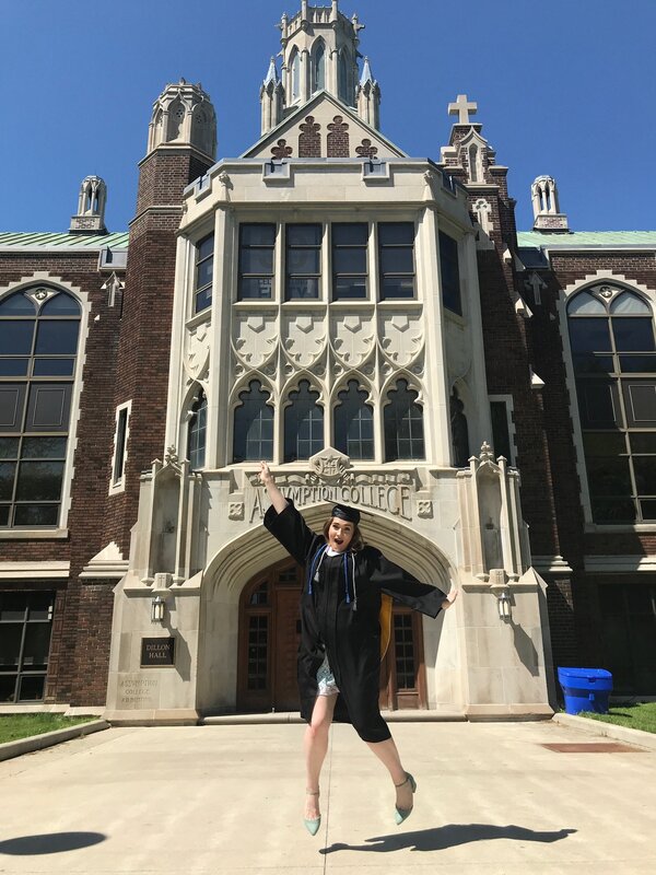 This is a picture of a woman in a graduation outfit jumping in the air in front of a school administration building.