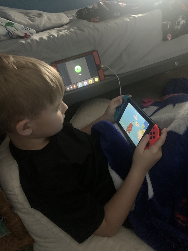 A child sitting next to a bed playing a Nintendo Switch while on a group video call.