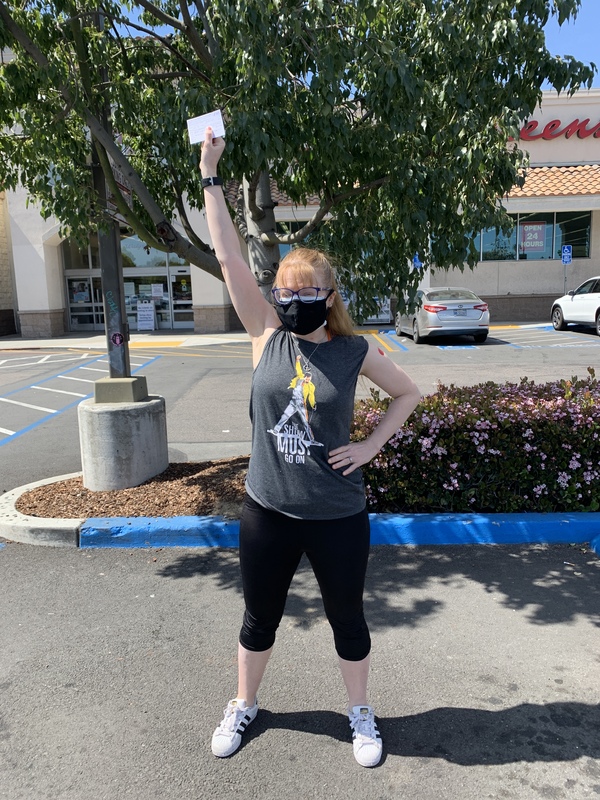 This is a picture of a woman wearing a grey tank top, black pants, glasses, and a face mask. She is standing in a parking lot holding what appears to be a vaccination card above her head.