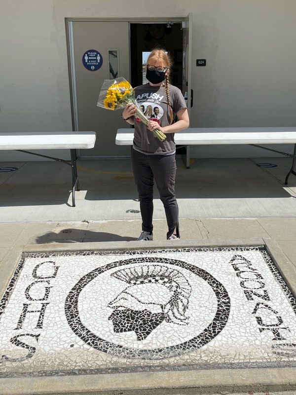 This is a picture of a woman carrying a bouquet of flowers in front of a few collapsible tables and an open door. She is wearing a black face mask, and an APUSH shirt.