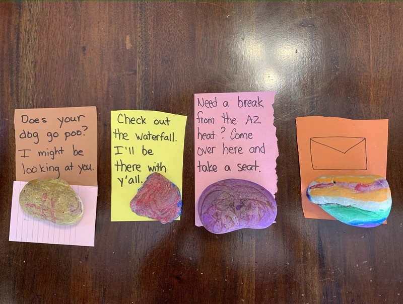 Four colorful rocks are sitting on pieces of paper that have clues written on them and are all placed on a wooden table.