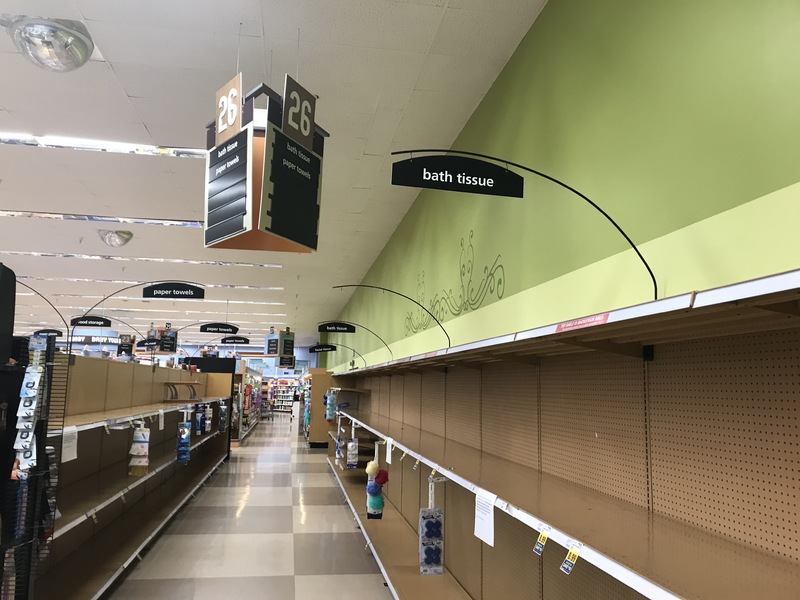 A supermarket that has green walls with checkered beige flooring. There are rows of empty shelves. Above the shelves is a black sign with white text that says "bath tissue". Hanging from the ceiling above the isle has a triangle shaped brown and beige sign that signs 26 with beige lettering.