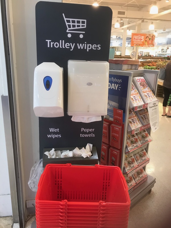 A black tolley wipe sign that has a dispenser for wet wipes and paper towels mounted onto it. Attached to the sign is a trashcan to dispose of the used wet wipes and paper towels. In front of the sign is stacked red baskets for customers.