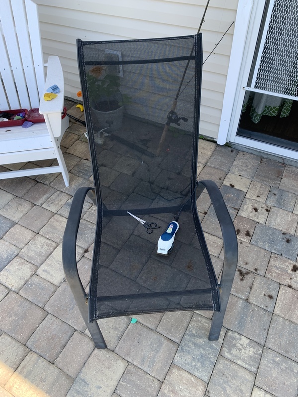 This is a picture of a black mesh patio chair sitting on a person's patio, with a pair of scissors and what looks to be an electric shaver resting on its seat. A fishing rod is leaned against the wall in the background, and another white patio chair with other objects on its seat is in the left hand corner of the photo.