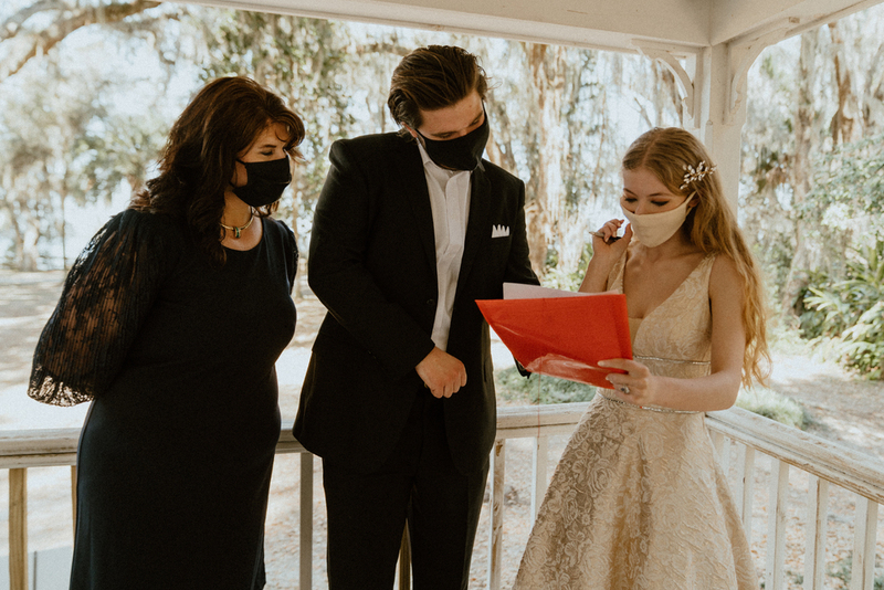 This is a picture of three people dressed in formal attire standing outside on a porch. All are wearing face masks. The woman in the photo seems to be wearing a wedding dress, while the man seems to be the groom.