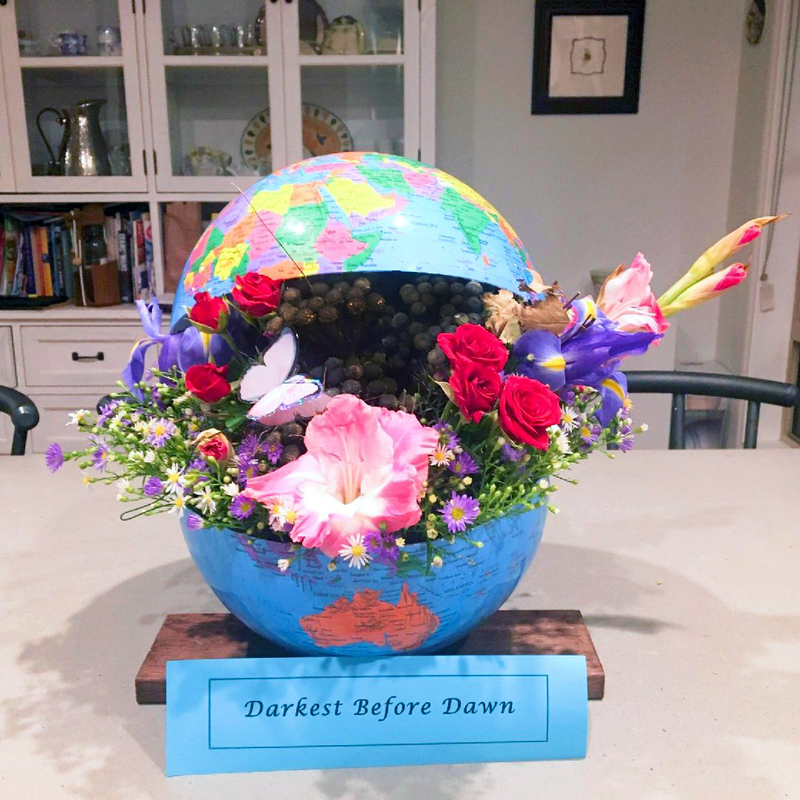 A globe is split in half with various kinds of flowers sticking out of it. The globe is sitting on a piece of wood with a piece of paper leaning against the front which says: Darkest Before Dawn. 