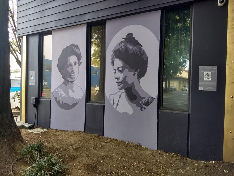 This is a picture taken of two murals that are painted on the side of a building. Two women in antiquated clothing are depicted in black and white.