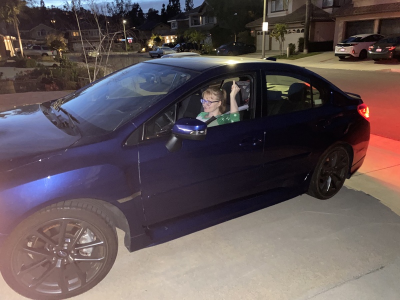 This is a picture of a woman in a green shirt and glasses smiling while raising her pinky finger in the air out of her car window in a driveway. The background of the photo is a residential neighborhood at night.