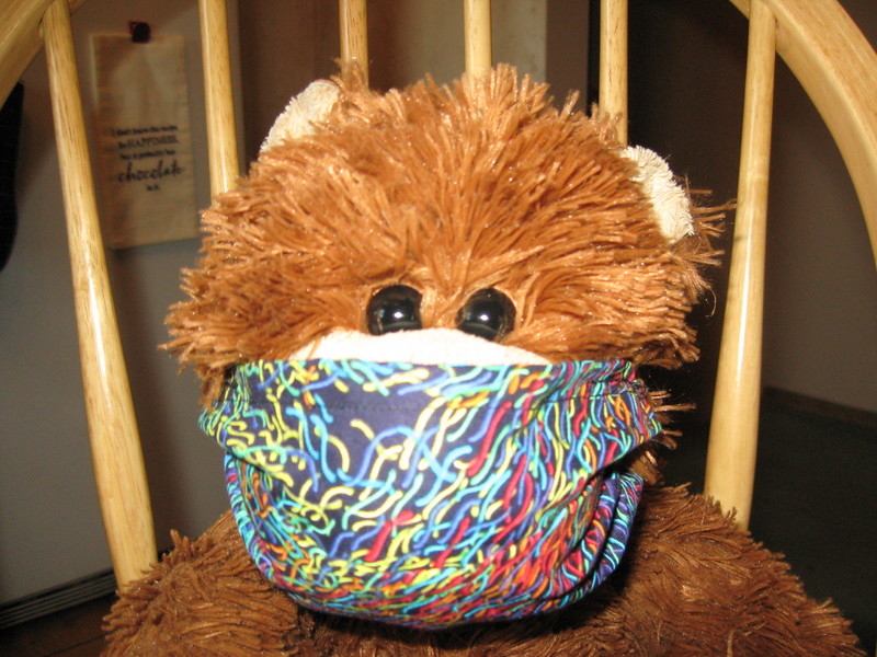 Teddy bear sitting in a wood chair wearing a multi-colored mask. 