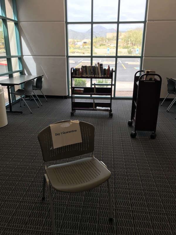 The corner of a library with windows. There are two book carts with books. There is a chair with a paper sign on it that says: Day 1 Quarantine.