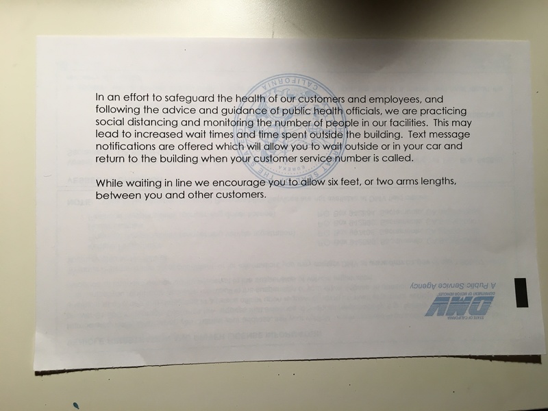 A paper information flyer from the Department of Motor Vehicles taped to a white wall saying they are practicing social distancing and limited number of people inside of their building.