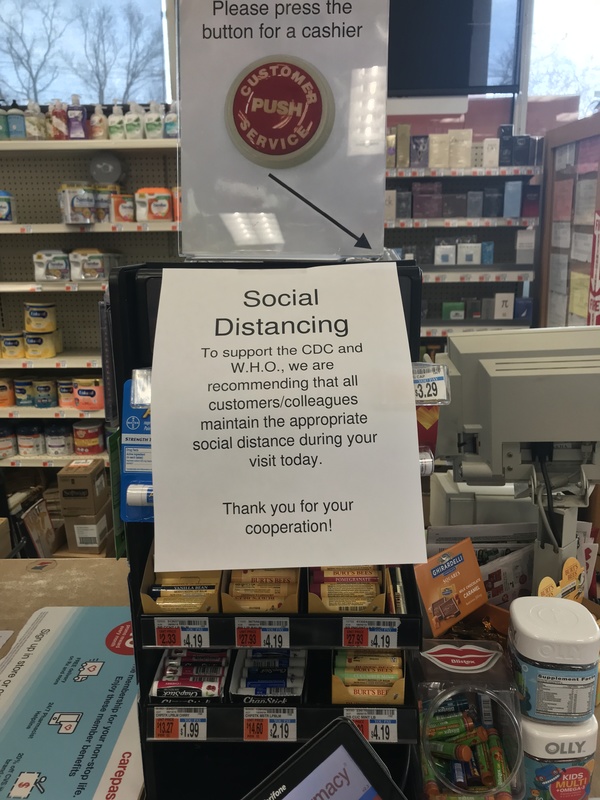 A register with a sign on it that says: Please press the button for a cashier and has an arrow pointing to the button. Below the sign is a paper sign that says: Social Distancing to support the CDC and W.H.O., we are recommending that all customers/colleagues maintain the appropriate social distance during your visit today. Thank you for your cooperation!
