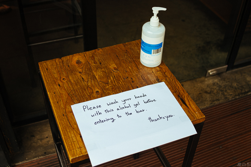 A wooden stool has a paper sign with written sharpie on it that says: Please wash your hands with this alcohol gel before entering the bar. Thanks you. There is a bottle of hand sanitizer on the right upper corner of the wooden stool.
