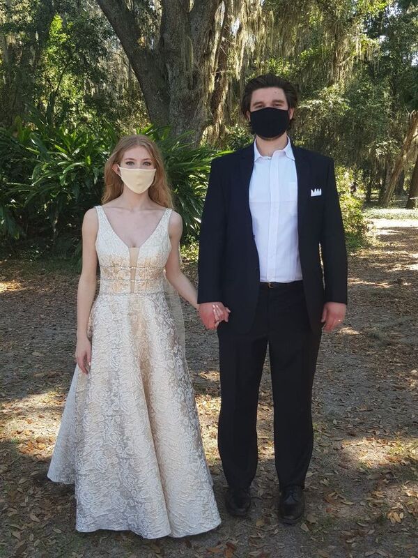 This is a picture of two people dressed in formal attire standing outside. Both are wearing face masks. They appear to be a bride and groom.
