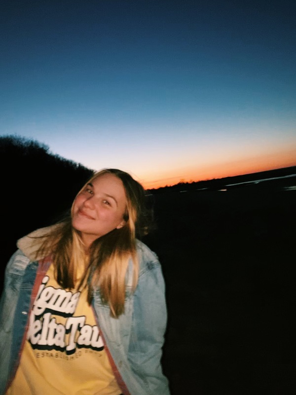 A person is smiling at the camera while the sunsets in the background.