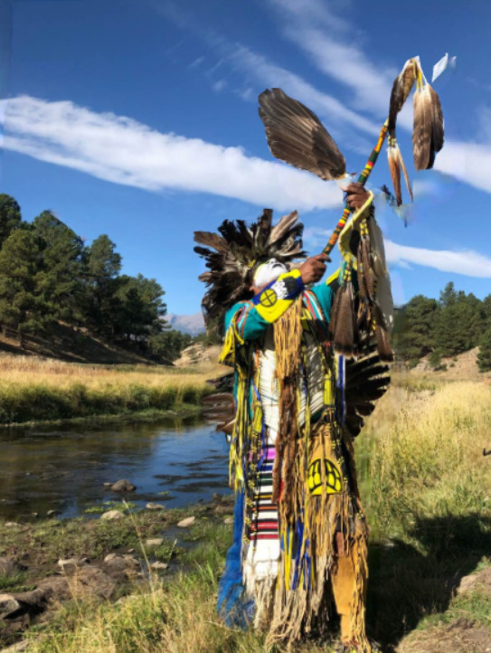 This is a picture of a Native American dressed in traditional ceremonial clothing outdoors by a stream, likely at a powwow.