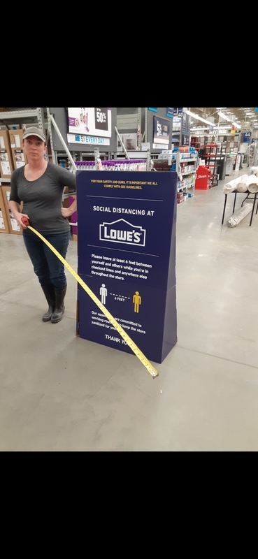 A person standing next to a sign at Lowe's talking about their social distancing policy while holding an extended tape measure.