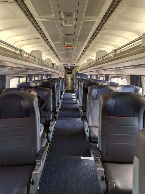 Inside of an Amtrak cabin that only has one person sitting in a seat off to the left. The rest of the cabin is empty. The isle has blue carpet to match the blue headrests of the seats.