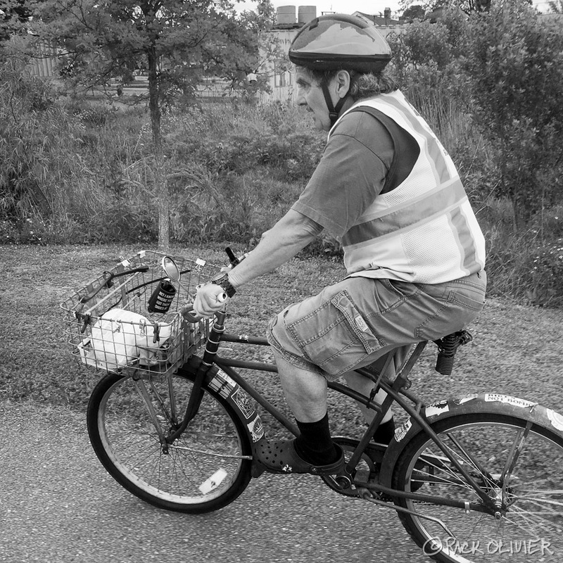 A man is riding a bicycle that is wearing a safety vest and bike helmet, on the front of the bike is a basket holding various items.