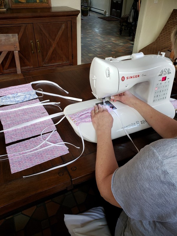 A person is sitting at a table using a white sewing machine sewing face masks. On the table off the left is scrap material for more face masks.