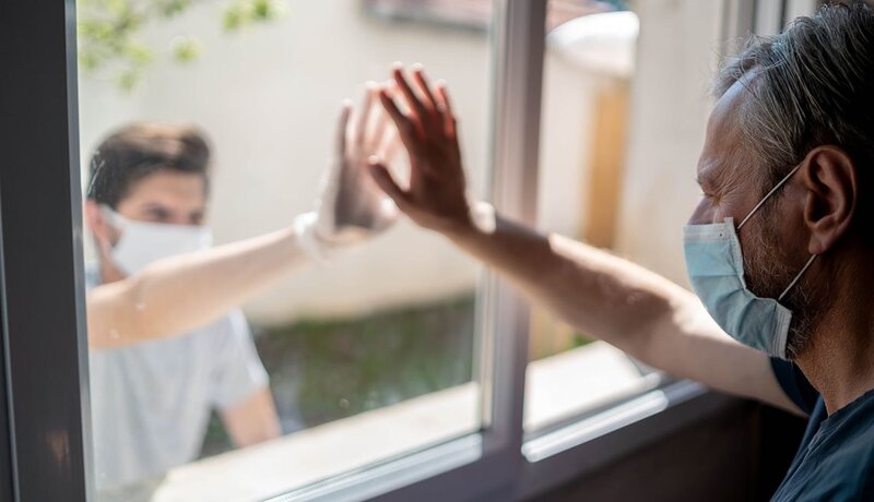 This is a picture taken of two people wearing face masks on either side of a window, and resting their hands on the same section of the window.