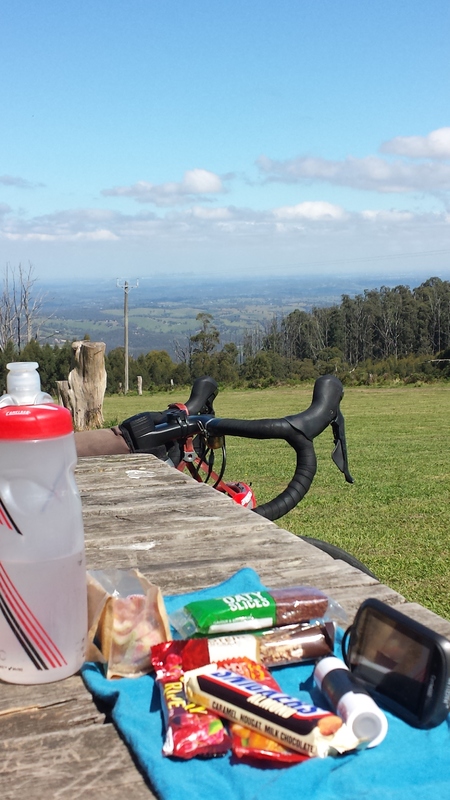 A picnic table that has a bike parked next to it. On top of the picnic table is a blue towel with various granola bars and a red water bottle.