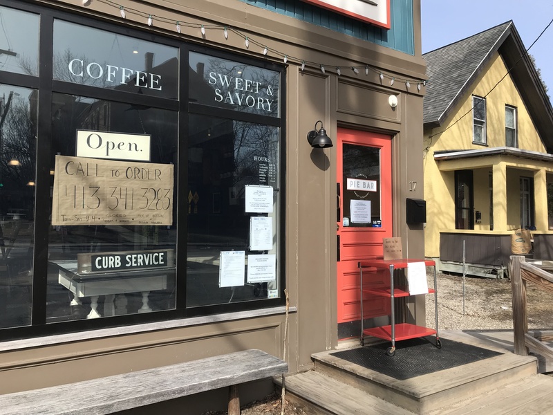 The front of a store has signs taped to the inside of the glass that says: Open. Call to order. Curb Service. In front of the orange door on the right is a red cart.