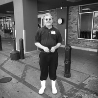 A man is wearing white rain boots with pants tucked into the boots. He is wearing a polo shirt with a name tag attached to it. The man is standing in front of a grocery store. The man is wearing circular sunglasses.