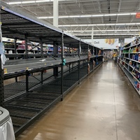 A supermarket is shown with white walls and brown concrete flooring. The empty isle has black and grey shelves on the right and on the left is white shelves that has various cleaning supplies.