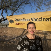 This is a picture taken of a smiling women posing in front of a yellow Arizona State University banner which reads "Operation Vaccination" on it.