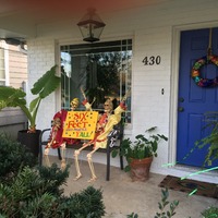 Front porch with Halloween decorations. Two skeletons hold sign that says, "Six Feet When Possible, Y'all!".