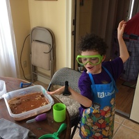 A kid with curly black hair is posing. He has an apron on over his purple shirt and he is wearing green goggles. In front of him is a brown wooden table with a square tupperware with brown water in it.