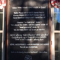 This is a picture of a sign outside a business which reads "Must wear Mardi Gras MASK to enter! Toilet paper SOLD OUT: Karen has it! Clorox wipes SOLD OUT: Karen has it! Hand sanitizer SOLD OUT: Karen has it! Cheese- 'to fold it in!': Aisle 9. 'Its about the WINE, not the label!': Aisle 12. 'Mall Pretzels!': Aisle 15. We do have SOAP so wash ya hands! Now offering 'Bebe's Chamber' while you shop. Curbside pick-up or home delivery available. 'Tweet us on Facebook'- Johnny Rose. LOVE, ACCEPTANCE, and LAUGHTER LIVE HERE! You are all ESSENTIAL. 'Frolic in the bawdy Dionysian bacchanal!'- Moira Rose." An image of a rose is present on the bottom of the sign.