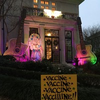 This is a picture of a house decorated with several COVID-19 related objects. A yellow sign stuck in the lawn out front reads "Vaccine, Vaccine, Vaccine, Vacciiiine!" Two giant acoustic guitars also flank the entrance to the home.
