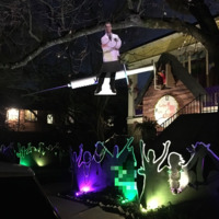 Exterior of home in New Orleans at night, lit with purple, green, and yellow lights. Cardboard cut outs of people raising hands and dancing are on the ground, and hanging above, suspended from a tree, is a cardboard cut out of a doctor.