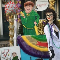 This is a picture of a series of decorations which encourage the viewer to make sure they get vaccinated. A character that resembles a monk clothed in a green robe is illustrated holding a needle and saying "Don't procrastinate, vaccinate!". Another mannequin dressed as a female doctor or nurse is next to this monk, and an object made to look like a COVID-19 particle stands on a pillar next to them.