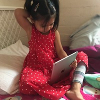 A girl with pigtails in a white and red polka dot dress is sitting on a bed with an iPad sitting in her lap.